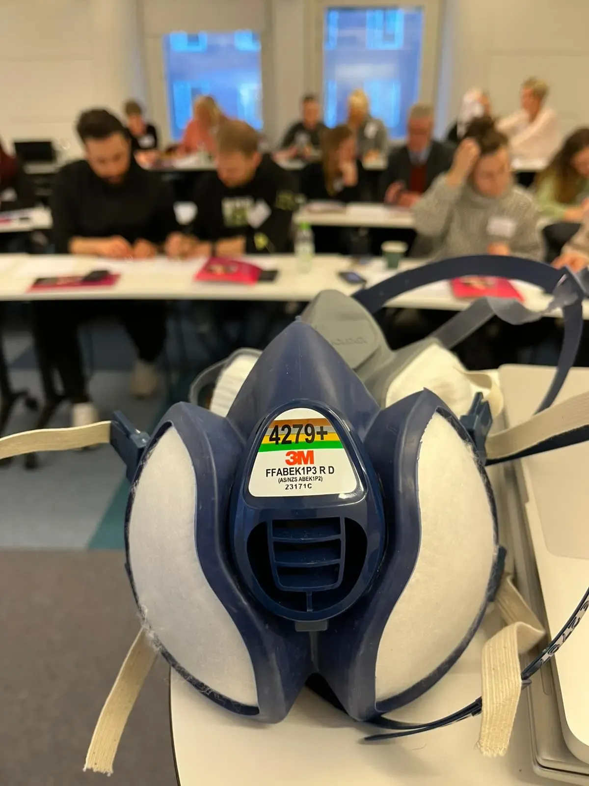 In the foreground, a PPE-maks, and in the background, out of focus, people sitting in a lecture hall