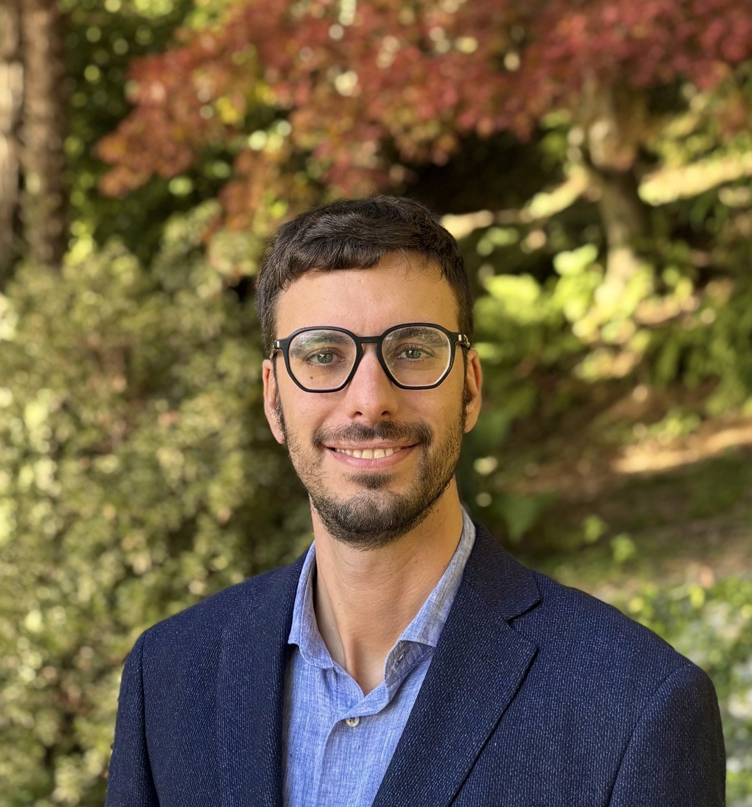Porträttbild av en man. Han har glasögon, blå skjorta och blå kavaj. I bakgrunden syns grön gräsmatta, träd och buskar.