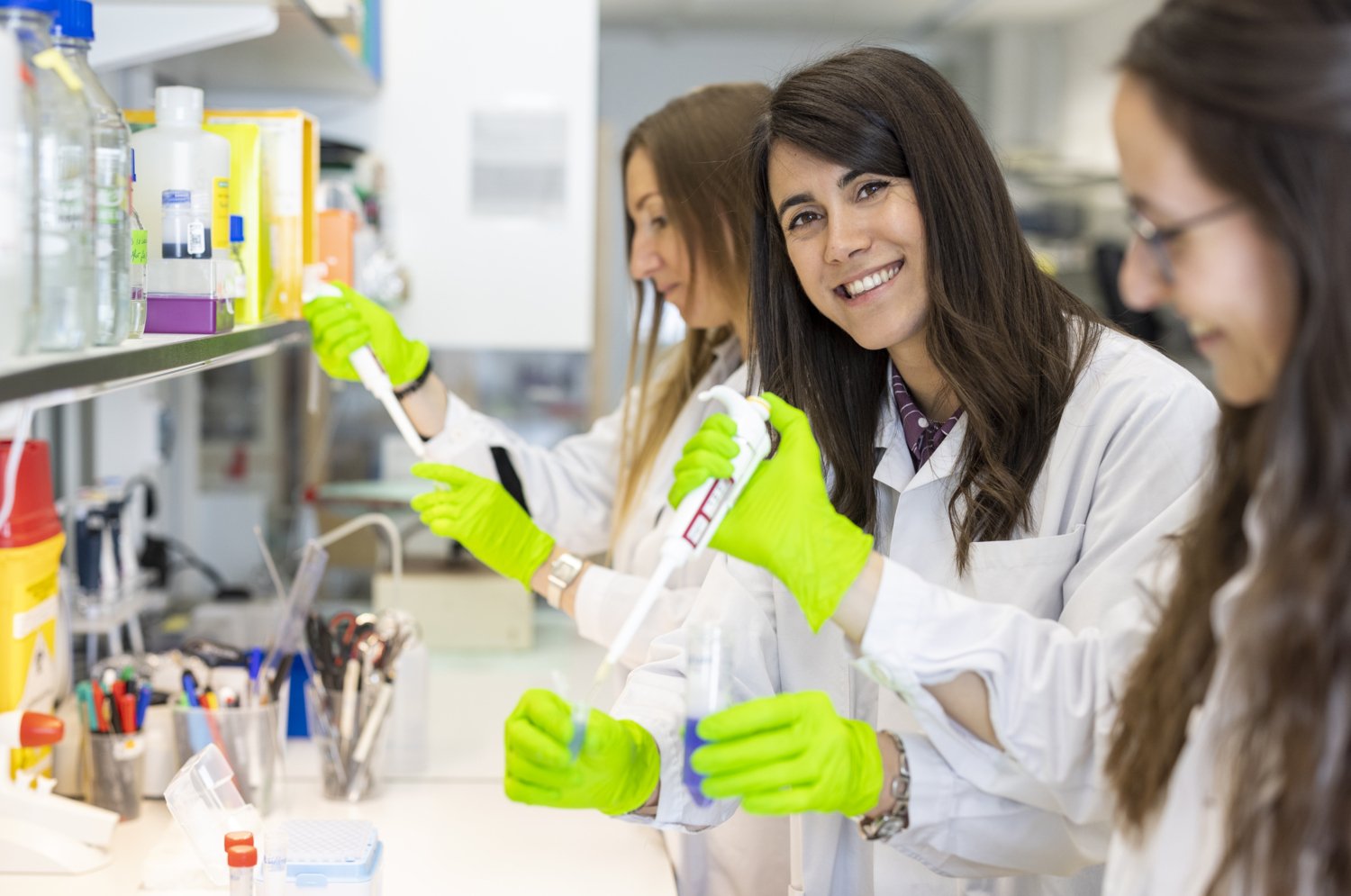 Tre kvinnliga forskare står med provrör och pipetter i labbmiljö. En av dem tittar in i kameran.