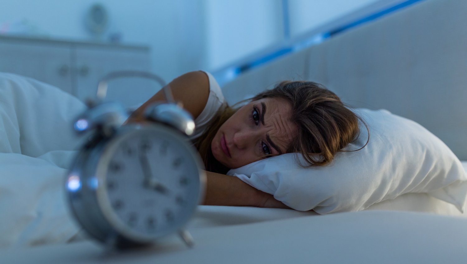 Woman lying in bed, frowning. In the foreground a clock with the hands pointing to four o'clock. There is morning light in the window above the bed.