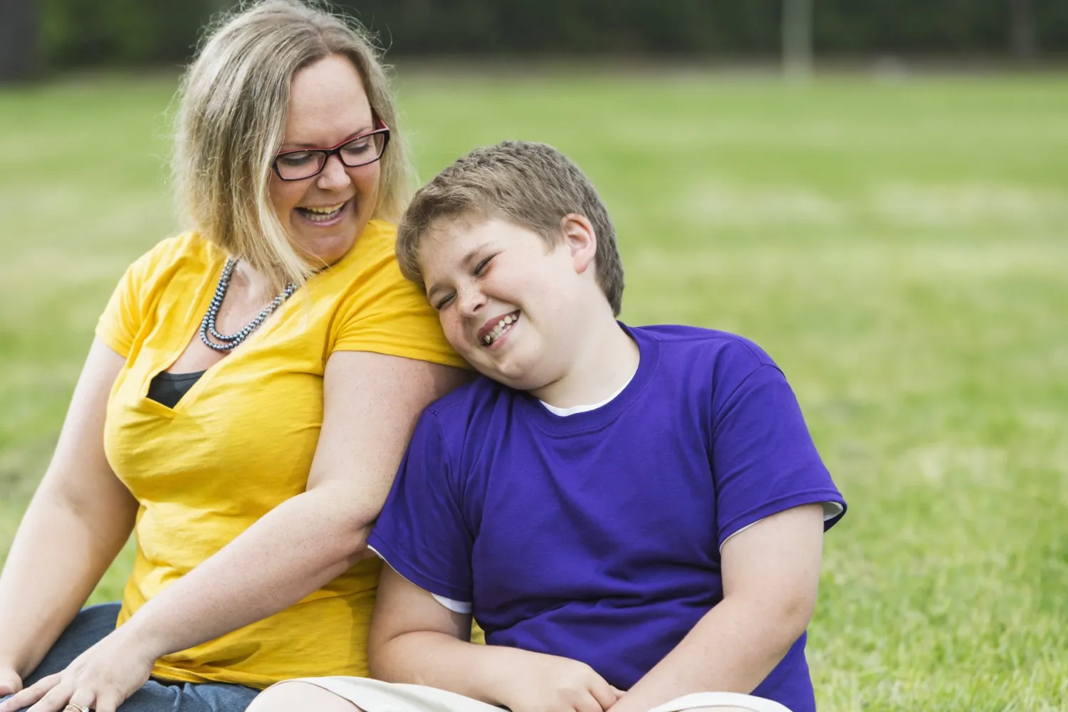 A mother and child, both overweight, are sitting on a sofa in a park. They are enjoying the sun, leaning against each other and laughing.
