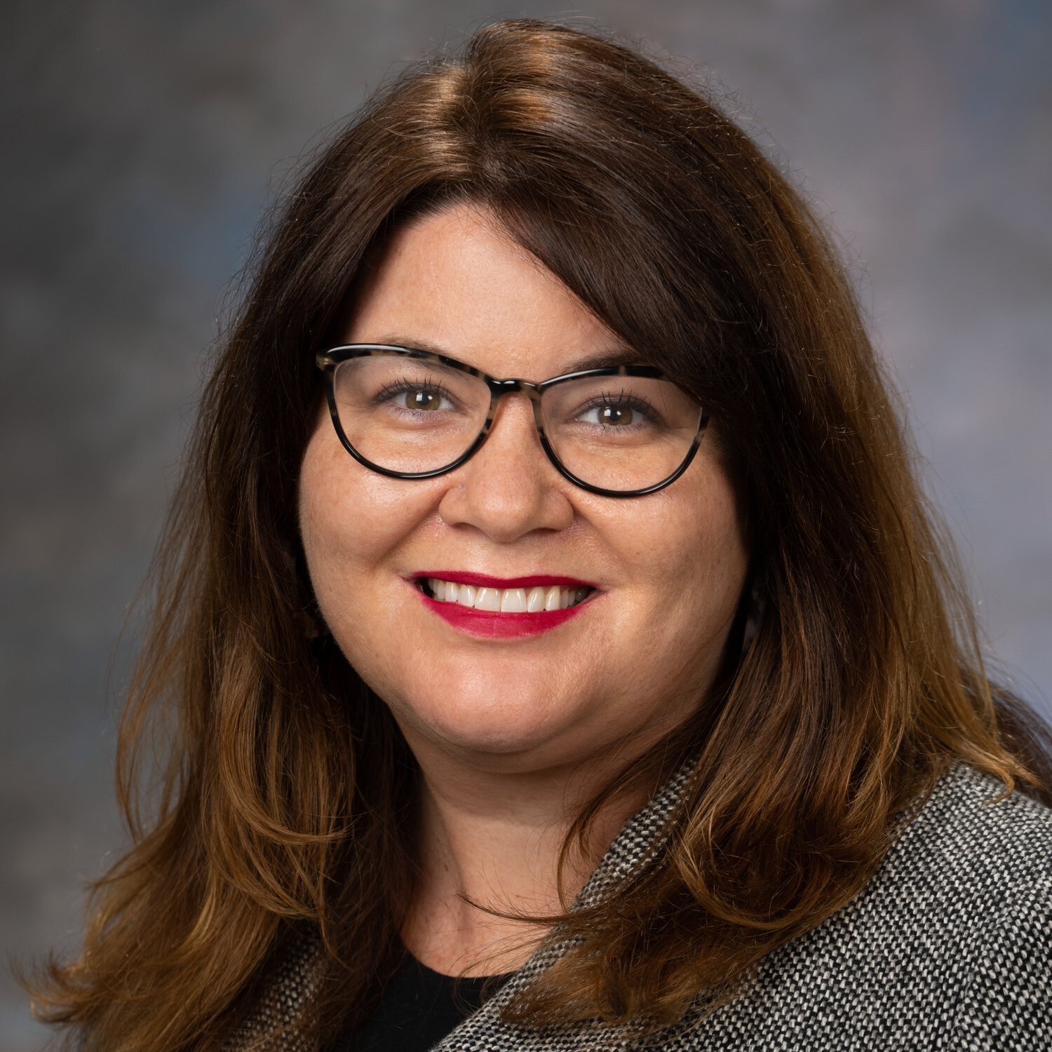 A woman with dark brown hair and glasses, wearing a knitted sweather, sitting in front of a dark grey background.