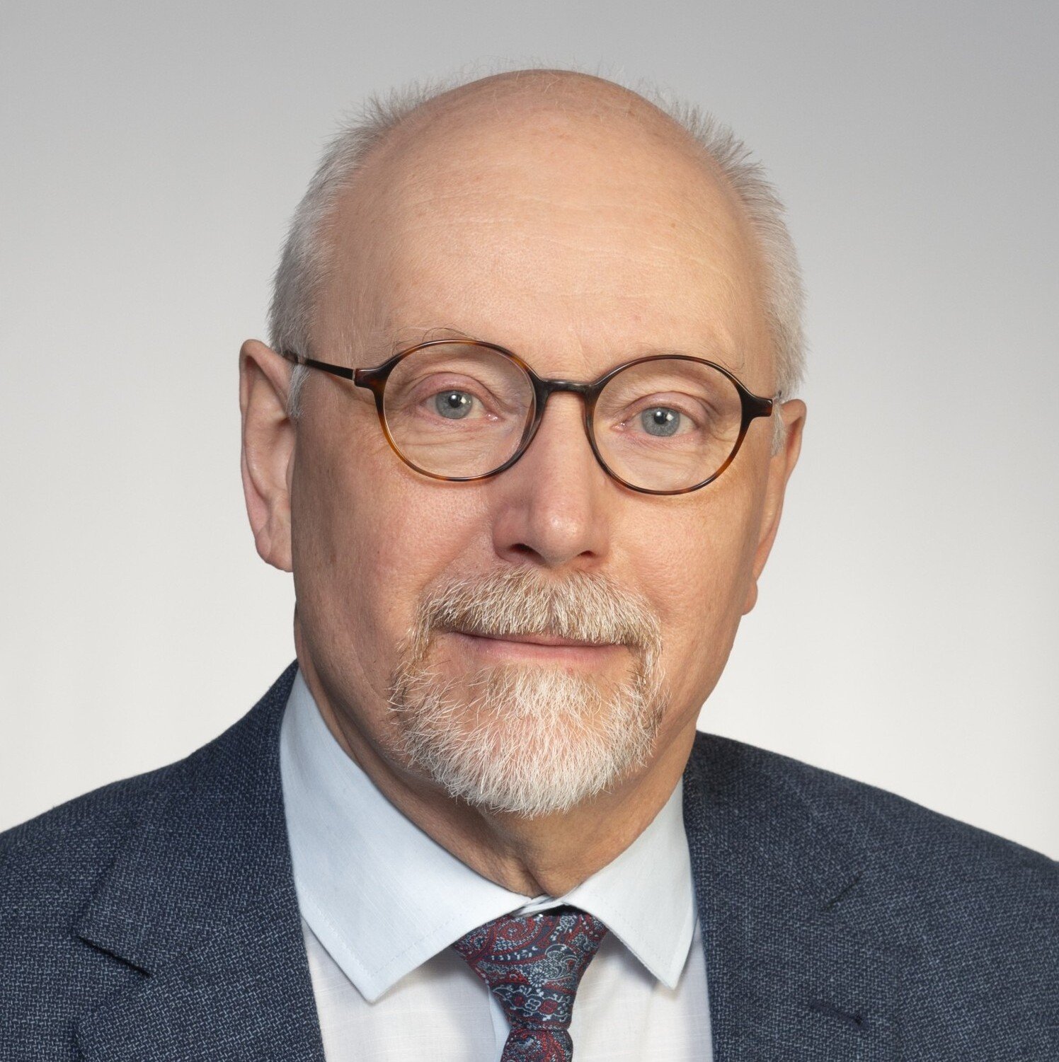 Porträtt av en man som bär kostym, slips och glasögon, som sitter mot en grå bakgrund.