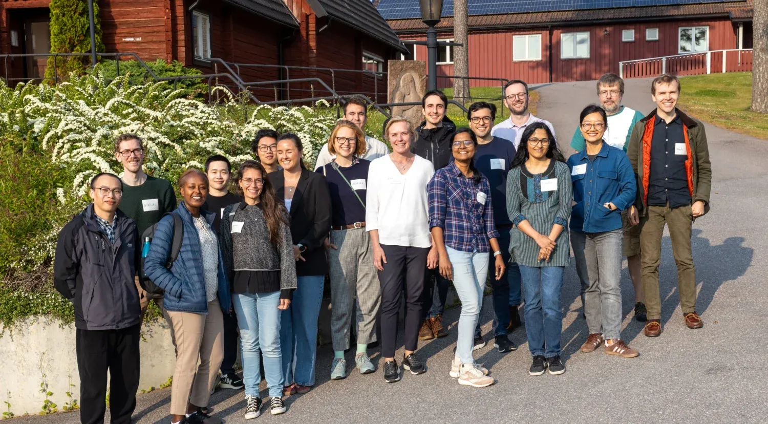 A group of people is standing outdoors in front of a building with red wooden walls and white window frames. Behind them are white flowering bushes, and the ground is paved with stone. The individuals are dressed in casual clothing and wearing name tags.