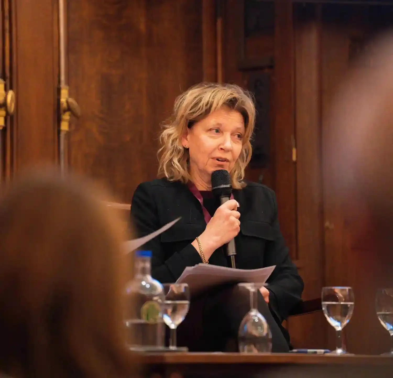 An audience is seated in a grand hall, facing a stage, where four people are seated with mics.
