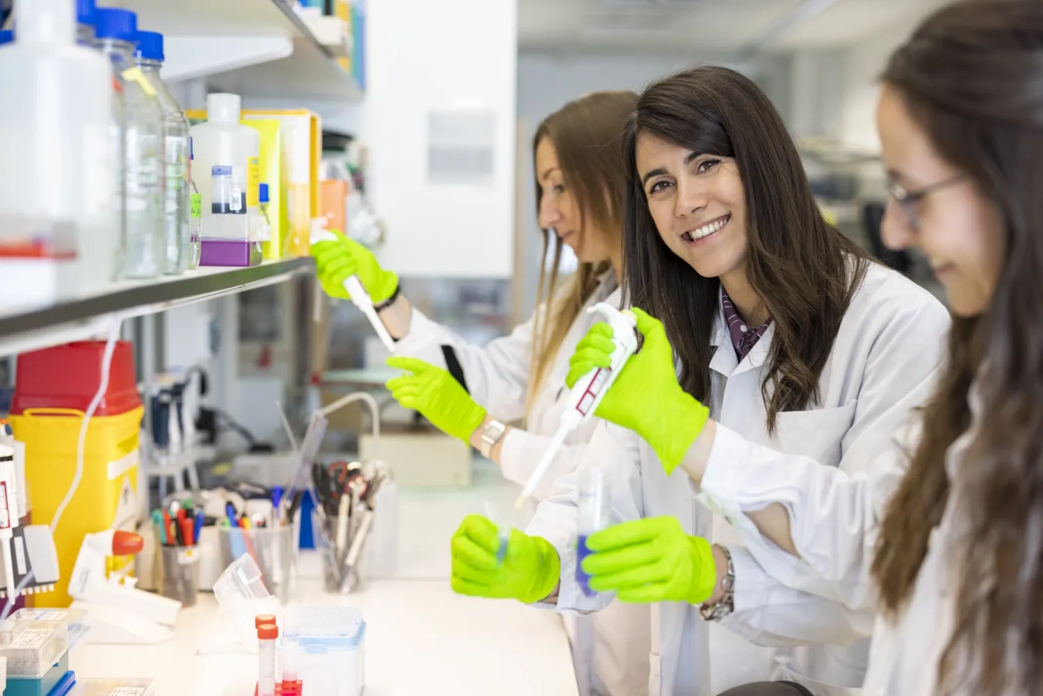 Three femare researchers standing with test tubes and pippettes in lab environment. One of them is looking in to the camera.