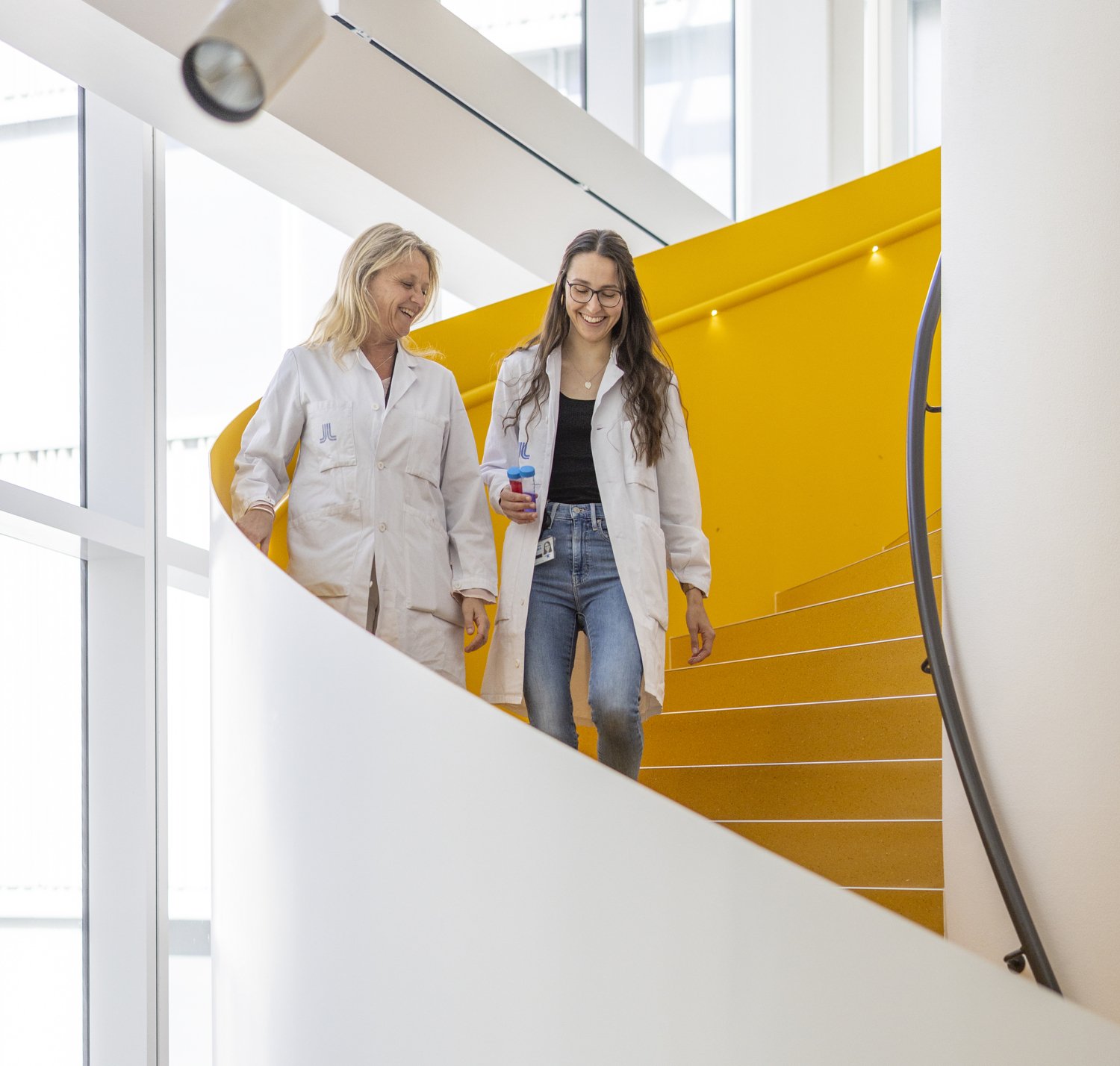 Two women in lab coats walking down yellow stairs.