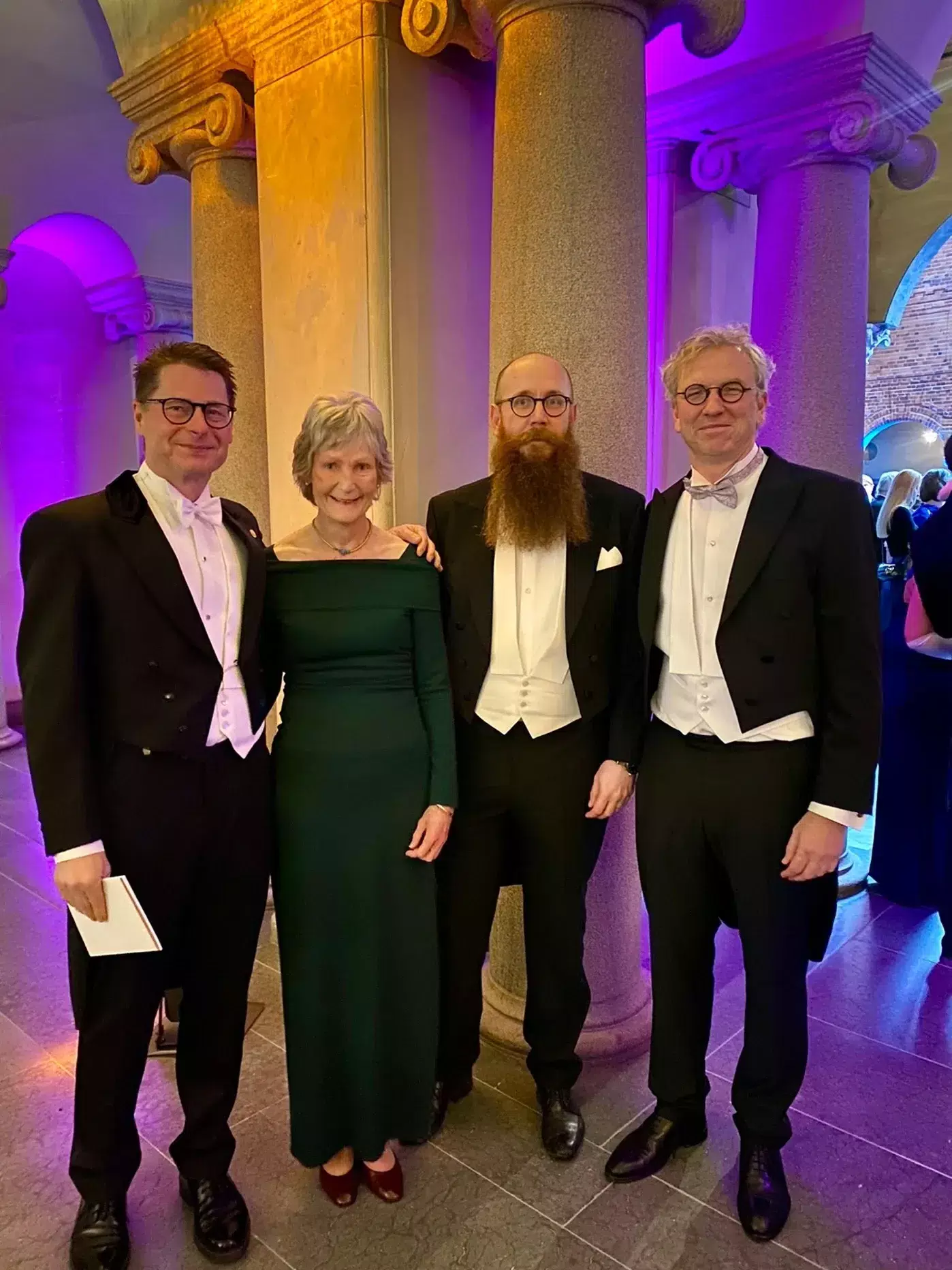 Three men and a woman are facing the camera and smiling. The setting is a room with pillars and purple lighting.