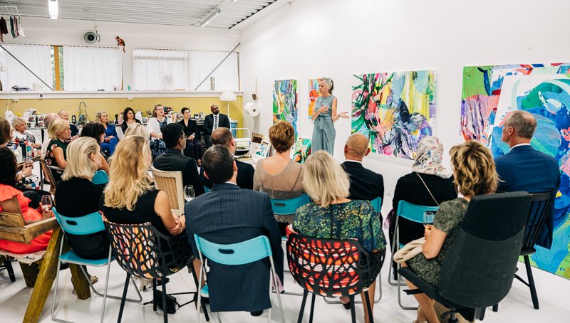 Group pf people listening to woman speaking, standing in front of paintings.