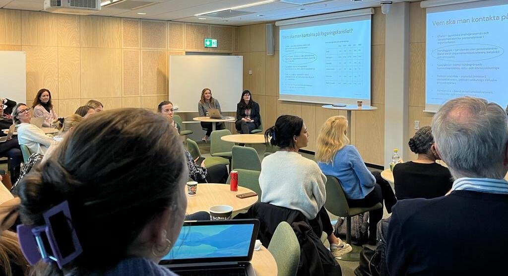 Ett rum med människor som sitter runt små bord och tittar framåt mot en skärm där det visas en powerpoint presentation