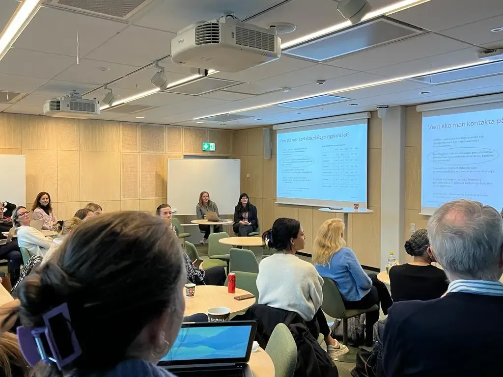 Ett rum med människor som sitter runt små bord och tittar framåt mot en skärm där det visas en powerpoint presentation