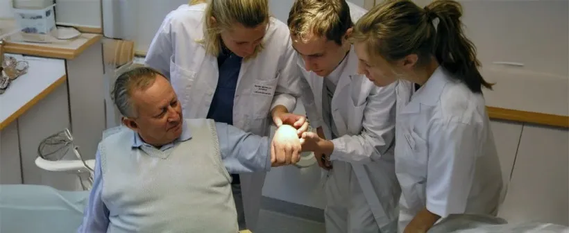 Three medical students stand around a patient and examine something on his hand.