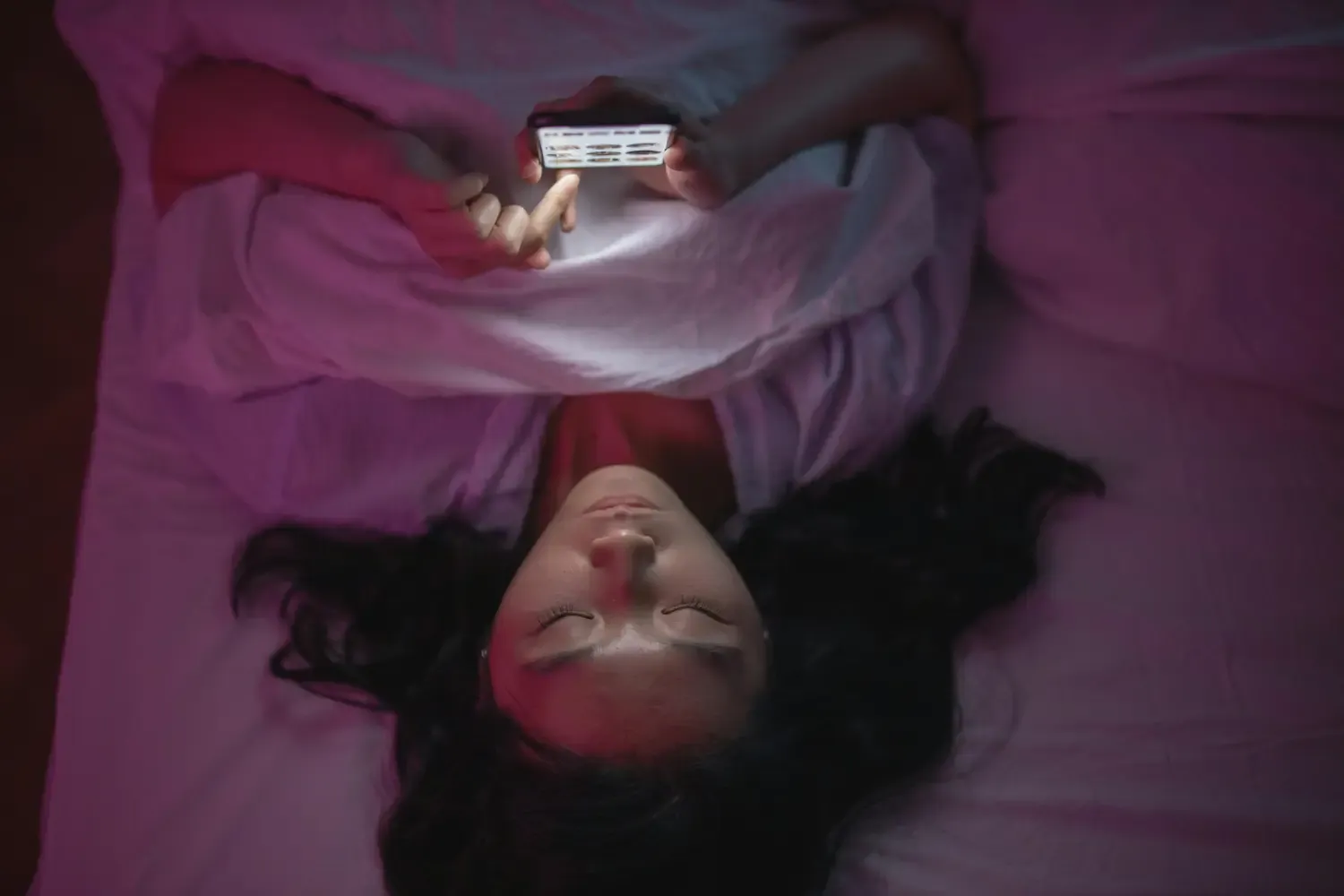 Young woman lying on her back in a bed lookaing at her phone. The light is dimmed.