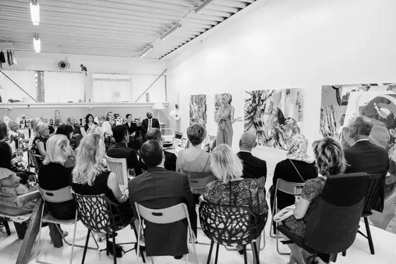 Group pf people listening to woman speaking, standing in front of paintings.