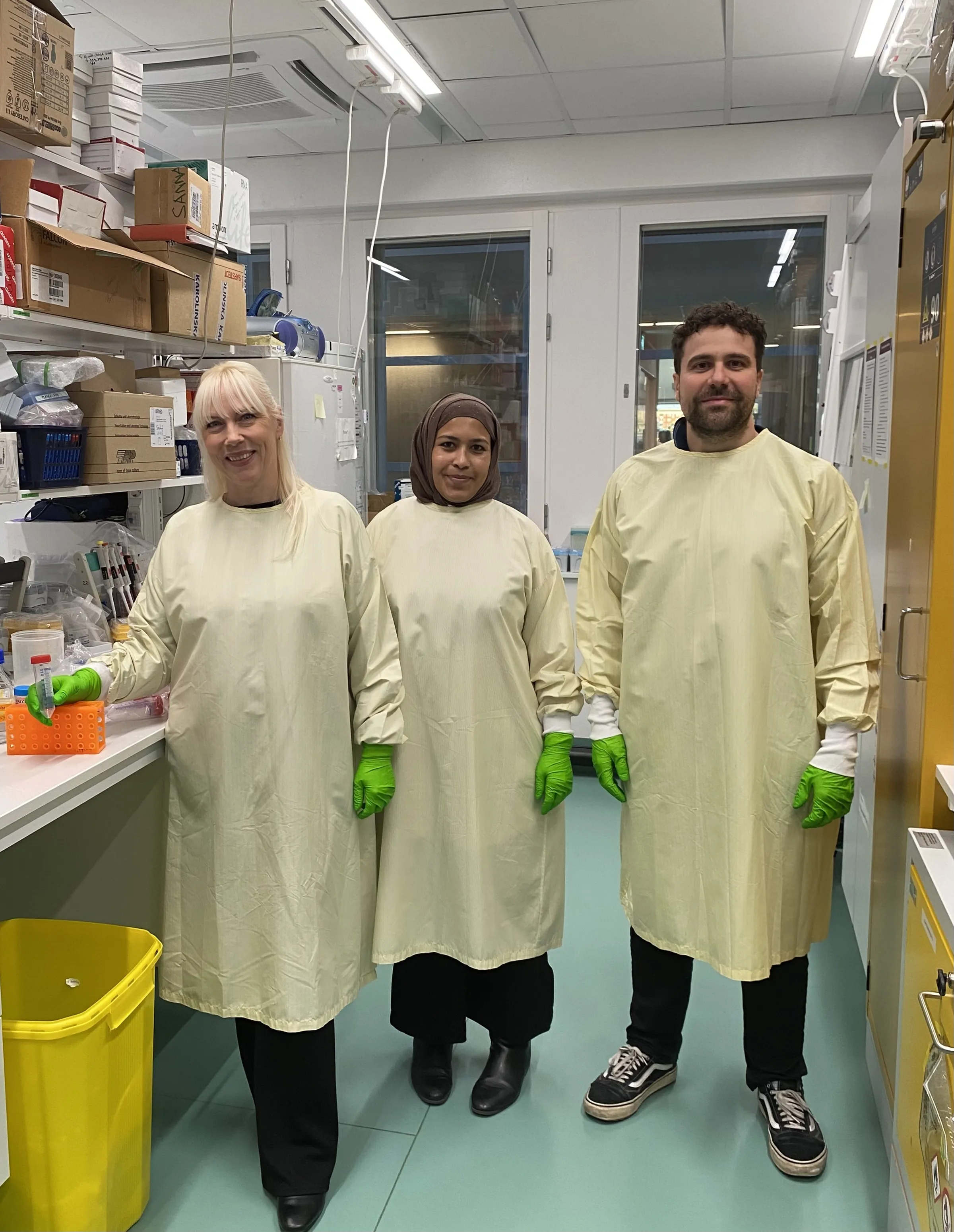 Three people are standing in a lab, dressed in lab coats and green rubber gloves.