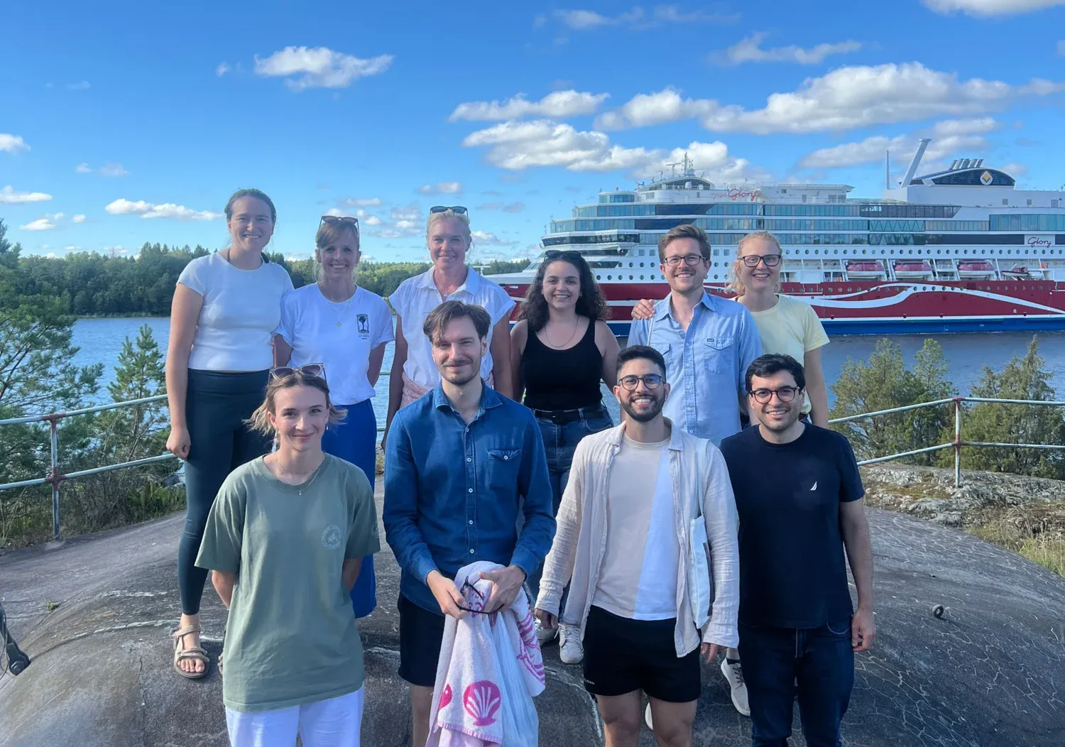 The group consists of 10 people standing on bedrock, and in the background, a large ferry is sailing on the water