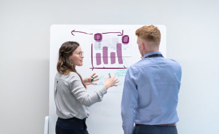 Two people standing in front of a white board discussing and writing.