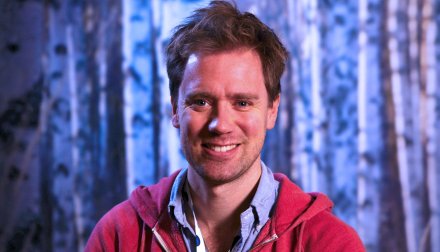 Färgporträtt på Eric Thelin som sitter framför en blå bakgrund i röd tröja.