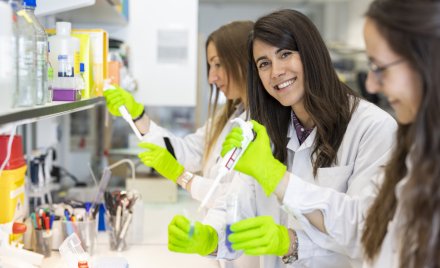 Three femare researchers standing with test tubes and pippettes in lab environment. One of them is looking in to the camera.