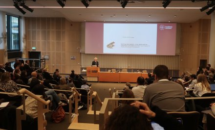 Photo taken from the back of a large lecture hall. At the front, a man is lecturing in front of a large projector screen.