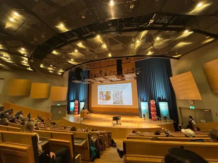 Photo taken from the back of a large lecture hall. At the front is a stage with a large screen and balloons on either side.