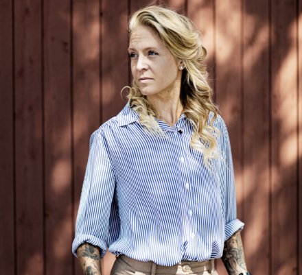 Portrait of Elin Mohlin standing against wooden wall outside.